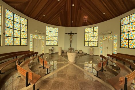 Interior da capela com vitrais coloridos e bancos de madeira em semicírculo
