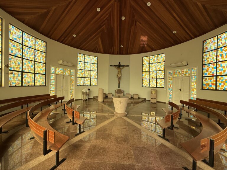 Interior da capela com vitrais coloridos e bancos de madeira em semicírculo
